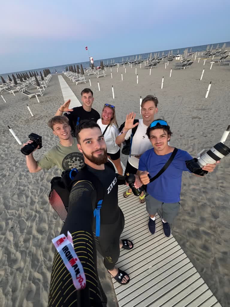 Štáb a závodník v neoprenu s páskou Ironman 70.3 si dělají společnou fotku na prázdné pláži za svítání.