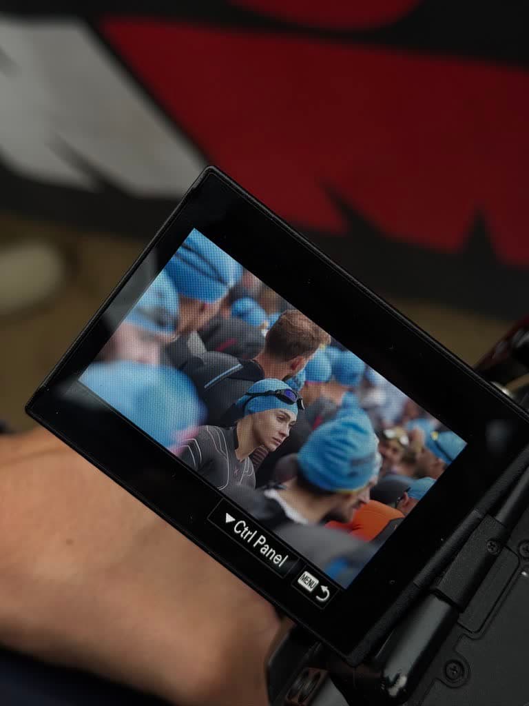 Displej kamery v ruce kameramana, na něm závodnice v modré čepici soustředěná před startem plavání.